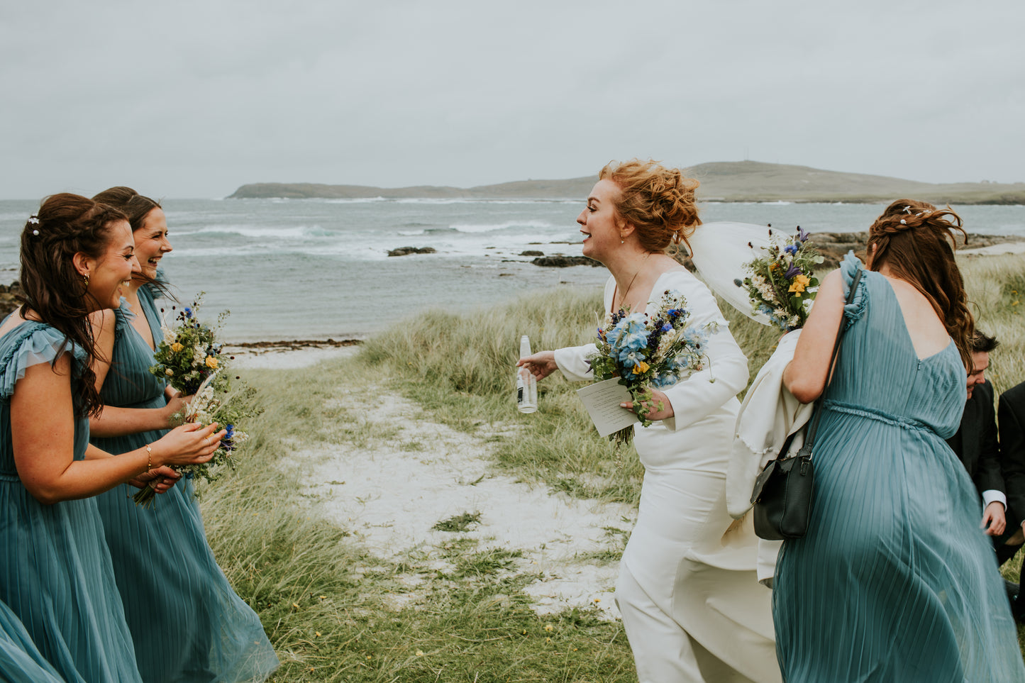 Woman in a white wedding dress with a bouquet, surrounded by bridesmaids in teal dresses by a coastal area.