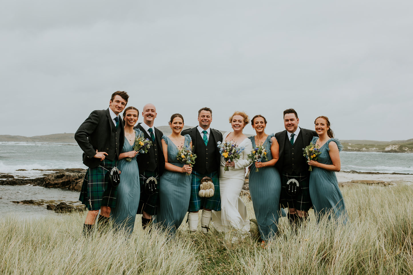 Group of people, including a bride and groom, standing in a grassy field with a body of water in the background.