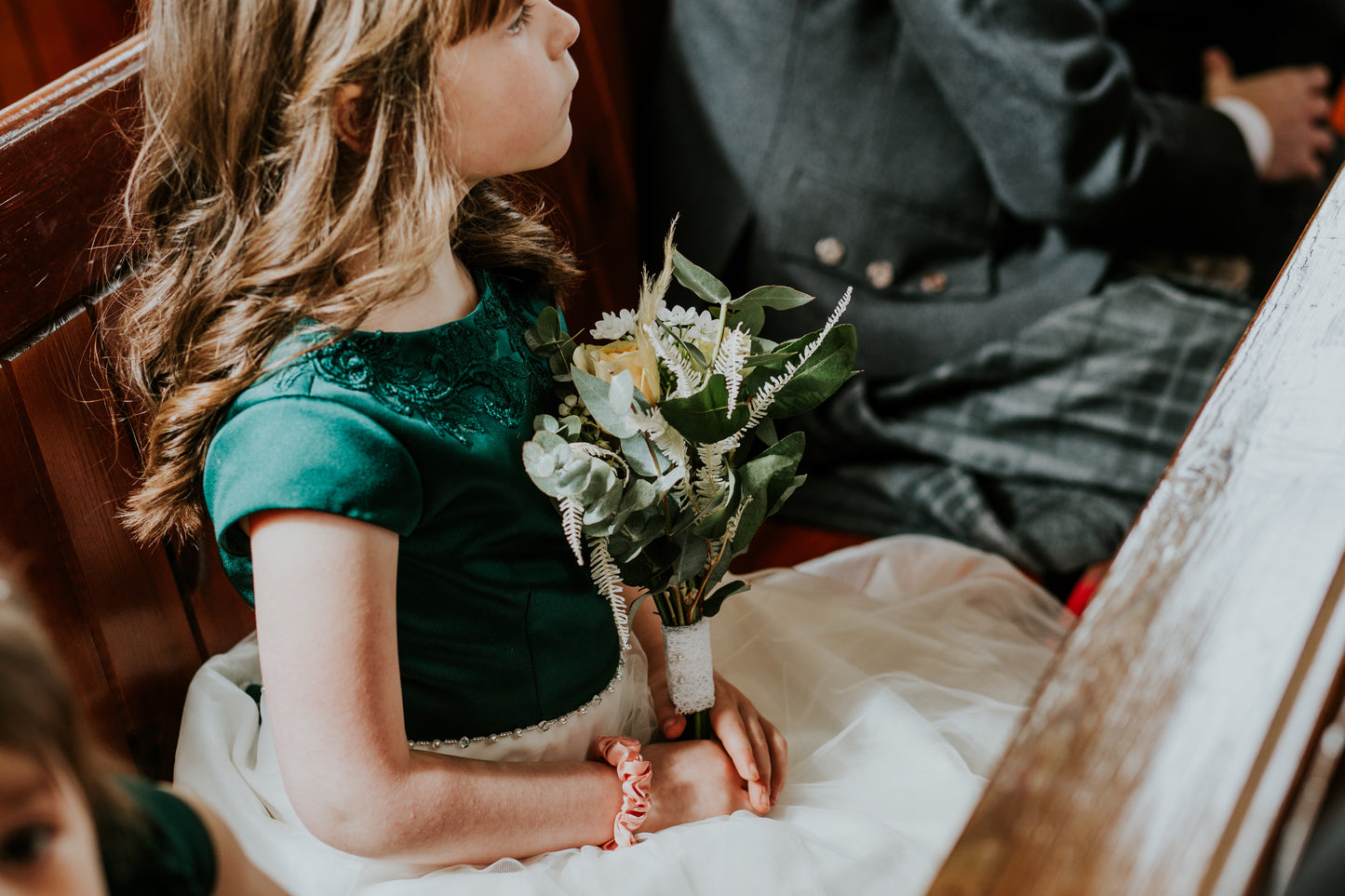 Young girl in a green dress holding a bouquet of flowers, sitting on a church pew.