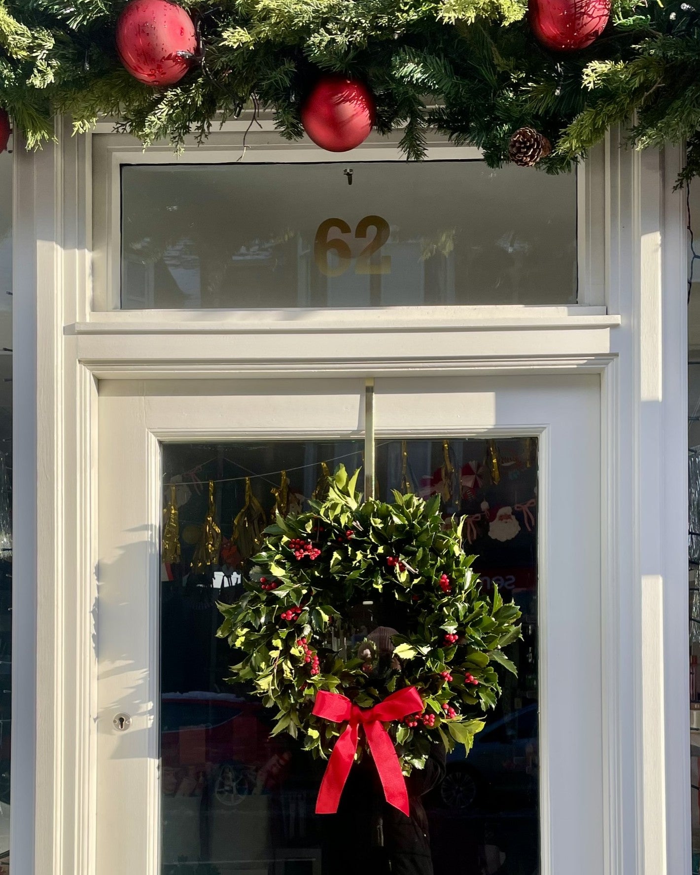 Decorative fresh holly wreath with red bow on a door with festive decorations above.