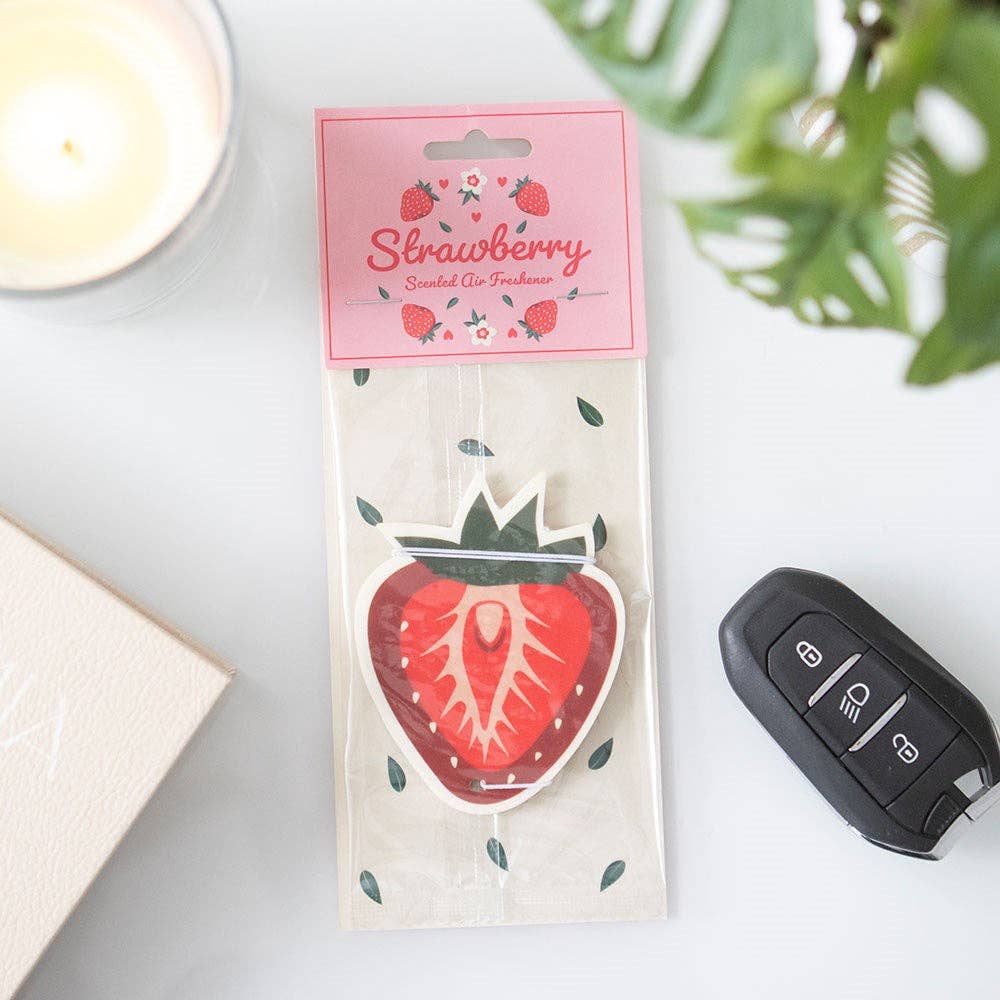 Strawberry-scented air freshener in packaging on a surface with a car key and candle.