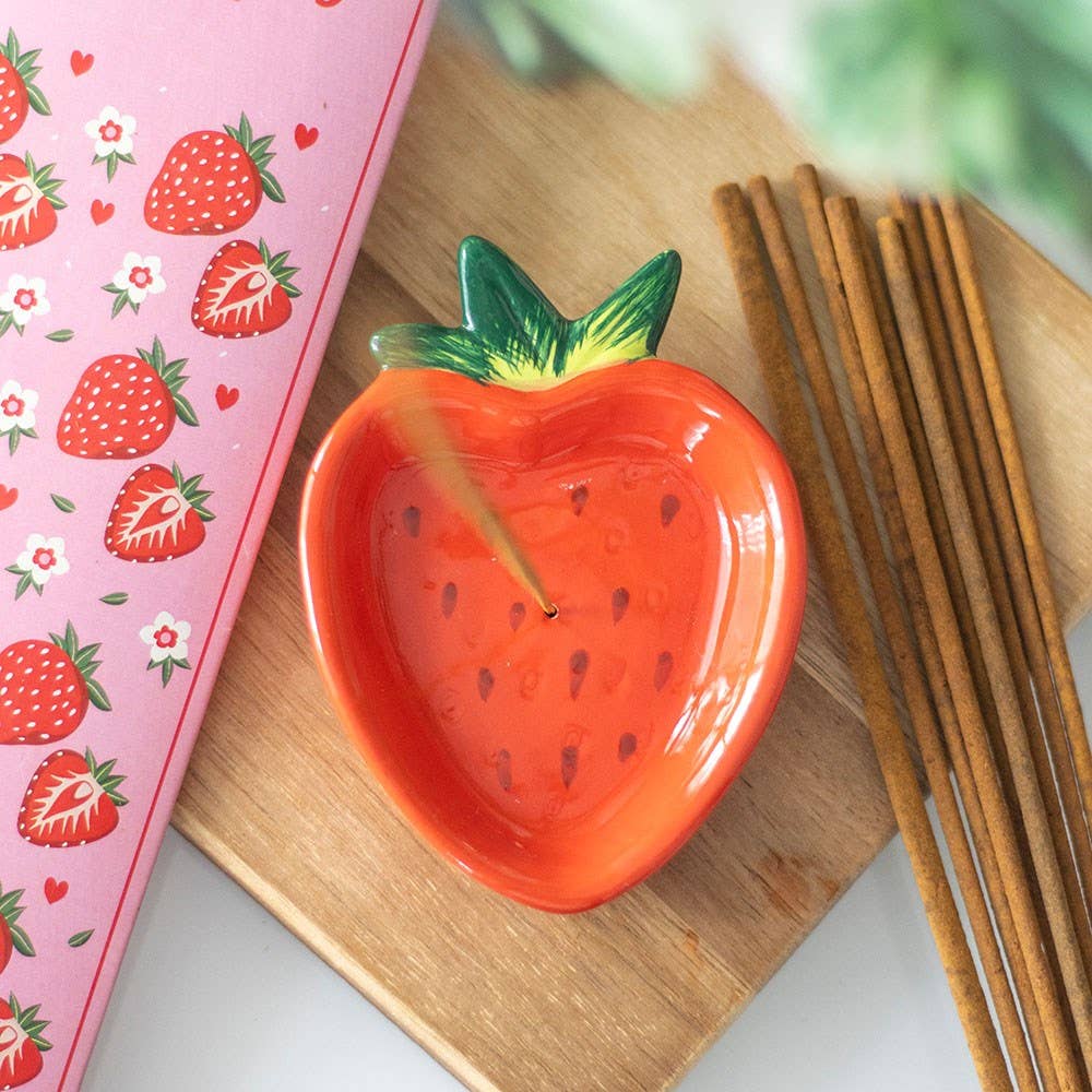Red strawberry-shaped dish with green leaves on a wooden surface next to a pink box with strawberry design.