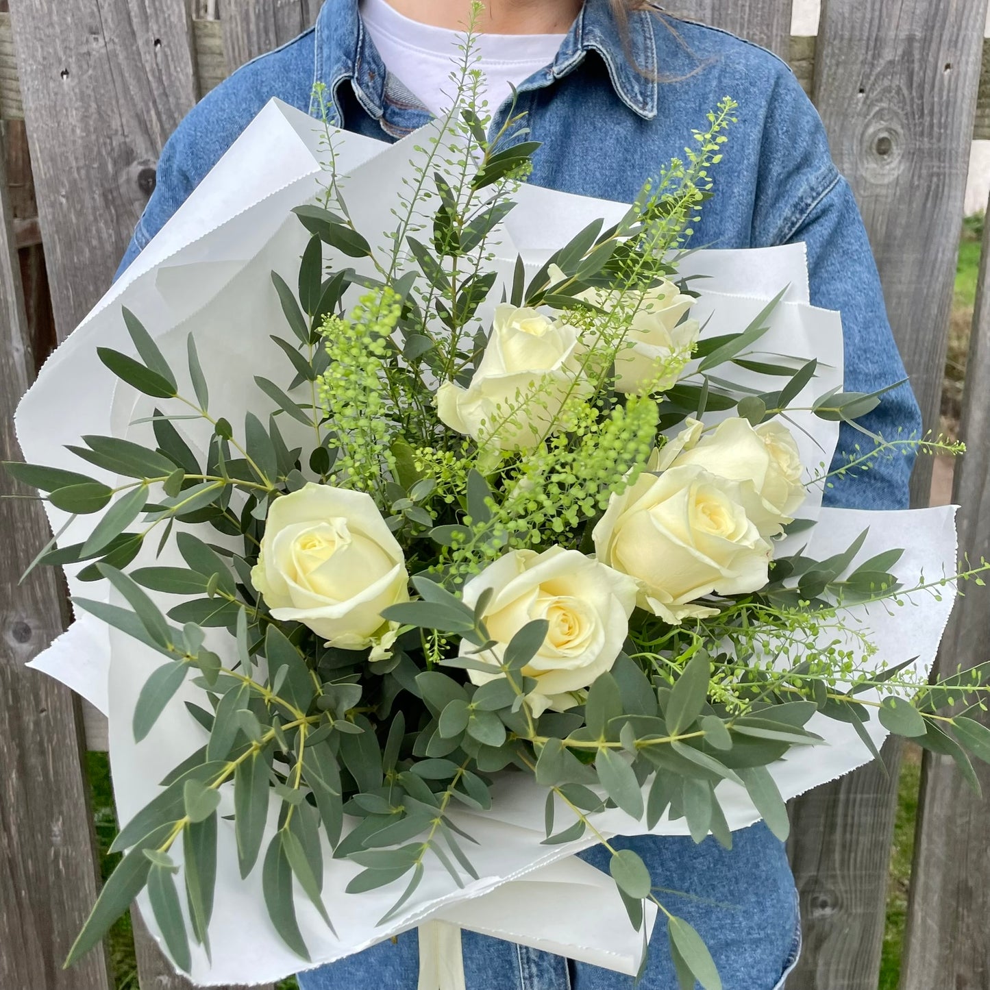 Bouquet of white roses with greenery held by a person wearing a denim jacket.